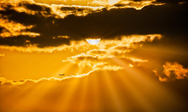 Silhouette of an airplane flying during a vibrant golden hour sunset with sunrays piercing the clouds.