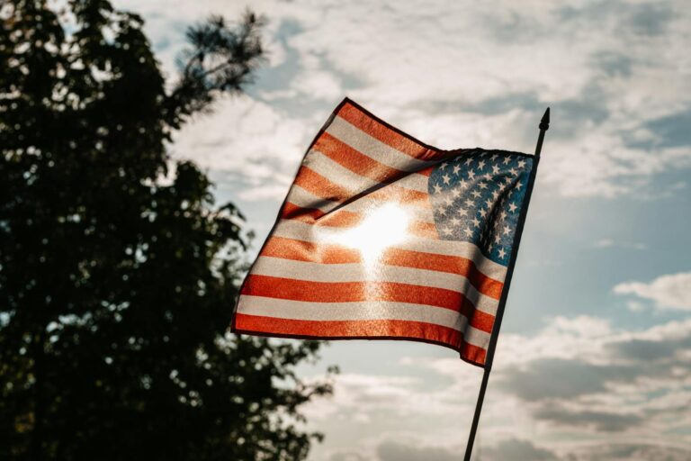 A waving American flag with sunlight streaming through against a backdrop of trees and cloudy sky.