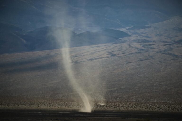 Aerial view of a dramatic dust devil swirling in a vast arid desert landscape under clear skies.