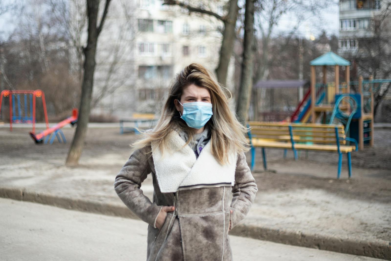 A woman in a coat and face mask stands in an urban playground, emphasizing safety.